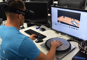 A person sitting wearing VR goggles, sitting in front of a computer, with their hand placed on metal plate. The screen of the computer is showing a virtual representation of the same screne.